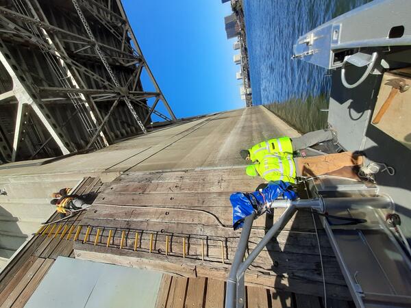 two people are high up on a bridge pier ledge while another crew assists from a boat parked close to the bridge pier. 