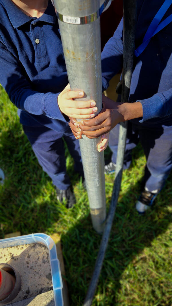 Two sets of children's hands holding onto a metal cylinder resting in the grass.