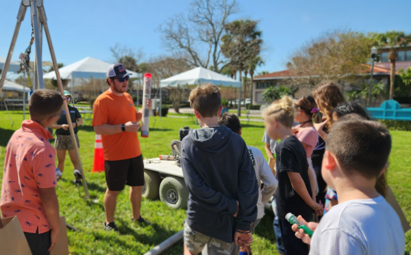 A USGS employee holds a display sediment core outside in front of a group of schoolchildren