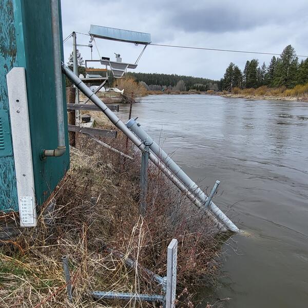 short steep river bank with dead grass. Equipment pipes in water next to grey aluminum cable car and green gage house