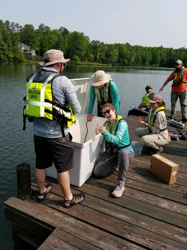 Six scientists in life vests gather and set up water quality monitoring equipment on a dock.