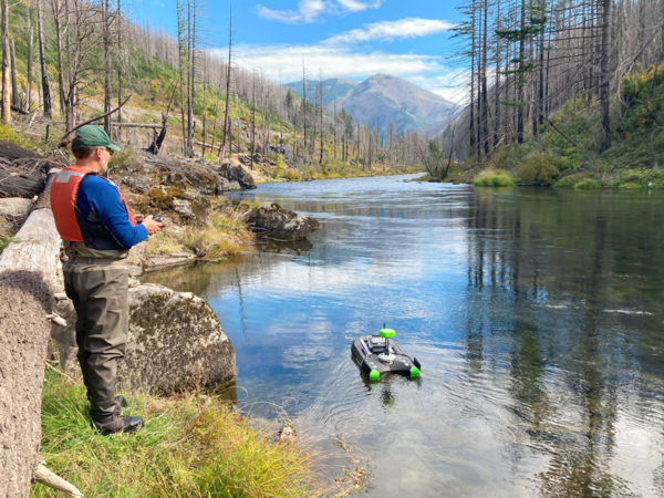 Male scientist operates a remote-controlled device from the riverbank in a burned forest landscape.