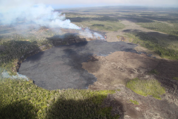 Helicopter view of a circular crater in the jungle, filled by black and brown lava. A lava waterfall glows on the far rim.