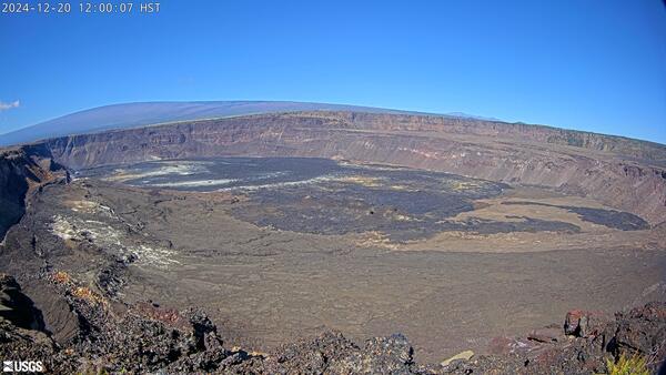 Desolate volcanic crater of gray/brown rick under a blue sky with a sloping mountain in the background