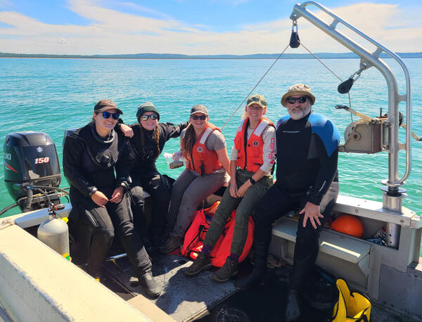 Five researchers posing on the deck of a small vessel on Lake Michigan. An engine and net sampling gear are present.