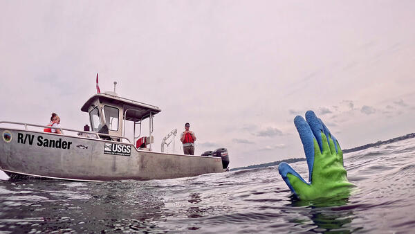 The gloved hand of a diver emerges from a lake and signals researchers on a small vessel, R/V Sander, in the background