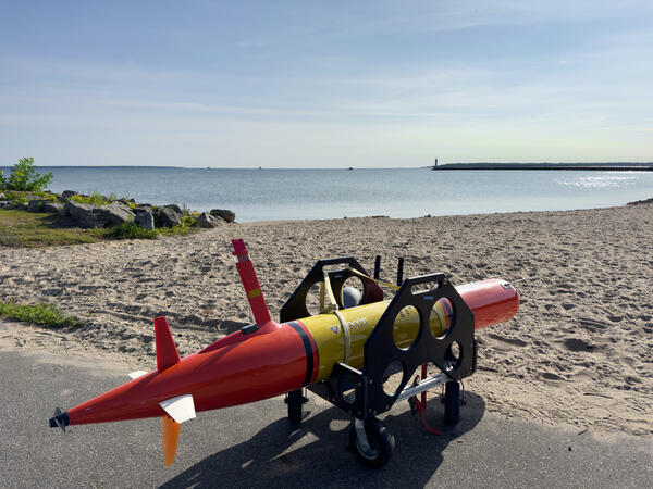 An orange Underwater Autonomous Vehicle in a transport cradle on a beach with a lake in the background.