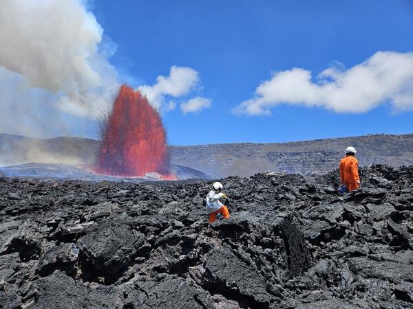 Color photograph of scientists on crater floor with lava fountain visible in the background
