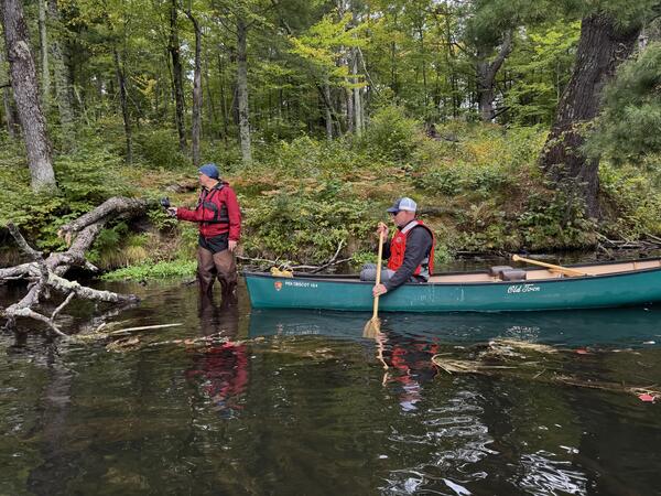 A man in a red vest stands in knee-deep water in a river with a handheld device while another man paddles a canoe.