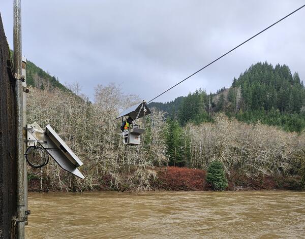 A man suspended above murky rushing river waters measures flow from an aluminum cable car