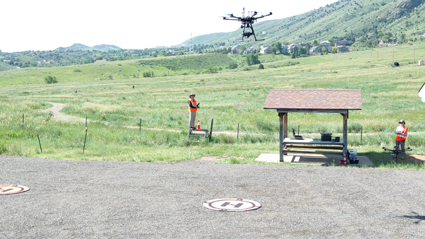 Pilots in a grassy field by a picnic shelter containing their instruments stand below a hovering drone