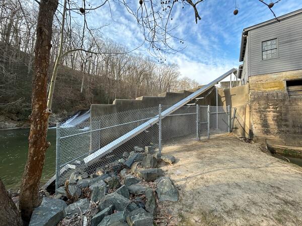 A concrete dam structure along a wooded river with a metal chute and chain‑link fencing beside a gray building.