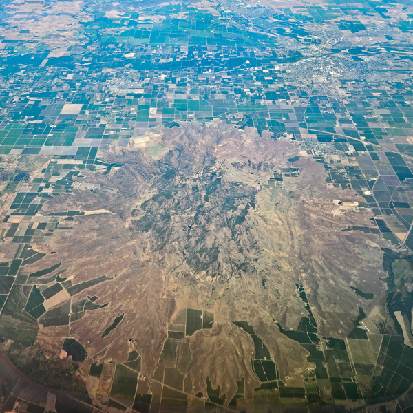A photo taken from an airplane at high altitude shows a cluster of craggy, brown and gray volcanic peaks surrounded by a circular apron of sloping, vegetated debris. Surrounded the volcano are green and brown agricultural fields laid out in a grid pattern.