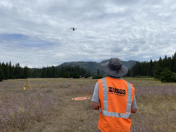 UAS pilot wearing USGS safety vest operates craft hovering above landing pad located in mountain clearing