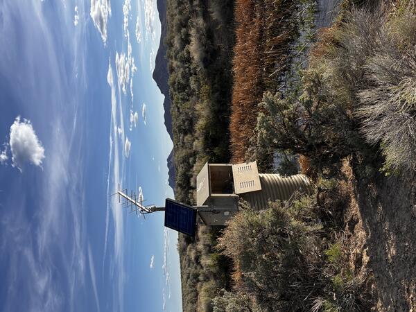 Tan solar-powered monitoring station in shrubland near ponding water with mountains under partly cloudy blue sky.