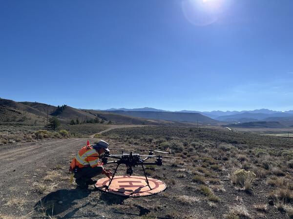 USGS drone pilot squatting by craft on landing pad located along dirt road with mountains in the distance.