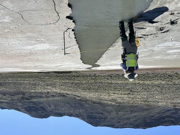 technician stands in ankle deep silty water running through the dry saline lakebed on a sunny spring day