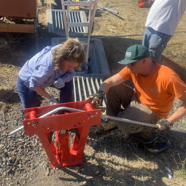 two people crouch near a pipe bender while feeding 1 inch metal pipe through