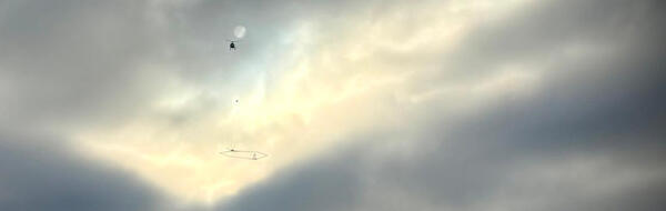 The silhouette of a helicopter with scientific instrument against a grey, cloudy sky 