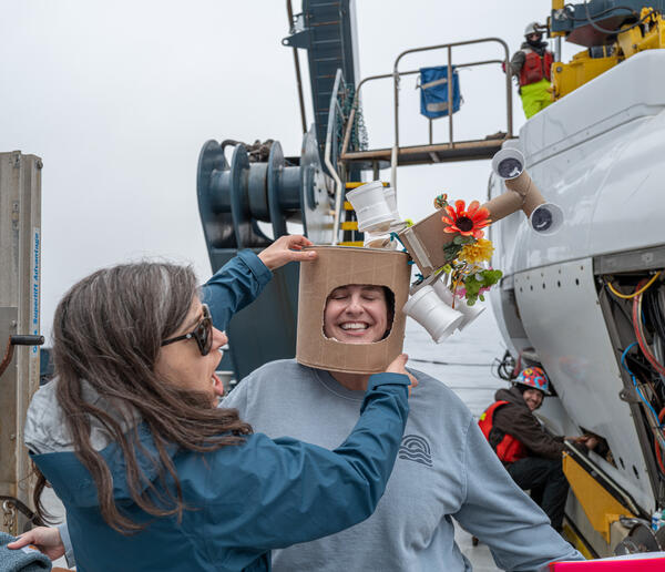 Two people on the deck of a boat laugh while one person wears a man-made hat