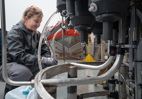 A person smiles while sitting on the deck of a boat as they collect water from tubes coming out of a oceanographic instrument