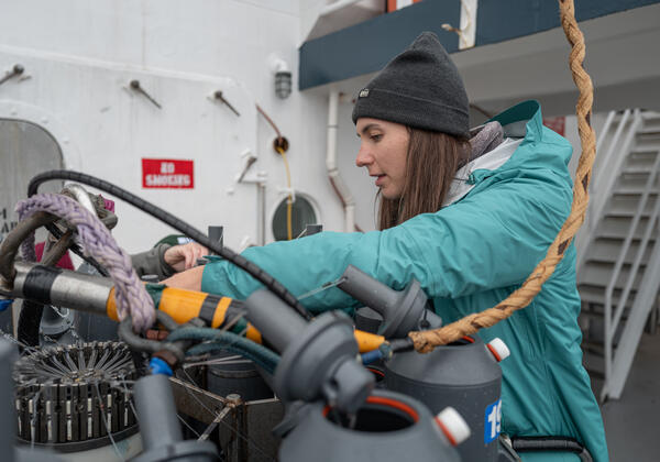 A person in a hat and teal jacket prepares Niskin bottles on a CTD