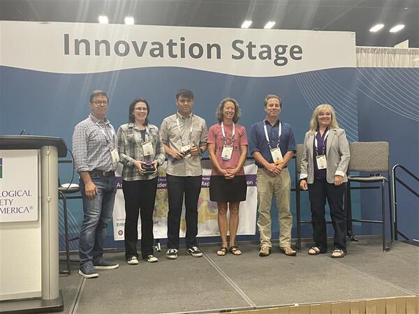 Photograph of six people on a stage with a blue background and high banner reading "Innovation Stage."