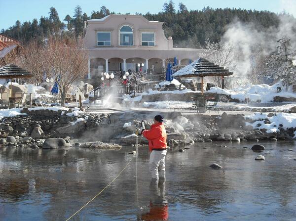 Female wearing orange float coat and black hat stands in calf deep water. Steam from hot springs visible.