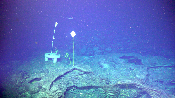 A white disk with three legs, bordered by several floating strings with flags, sits on a volcanic rock outcrop at the bottom of the deep ocean. The surrounding water is deep blue and murky.