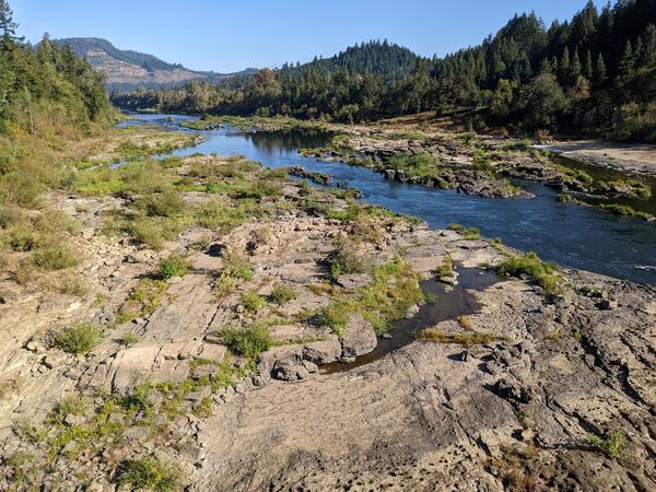 a wide, rocky riverbed with shallow, clear water in the foreground. Rolling forested hills and blue sky in background
