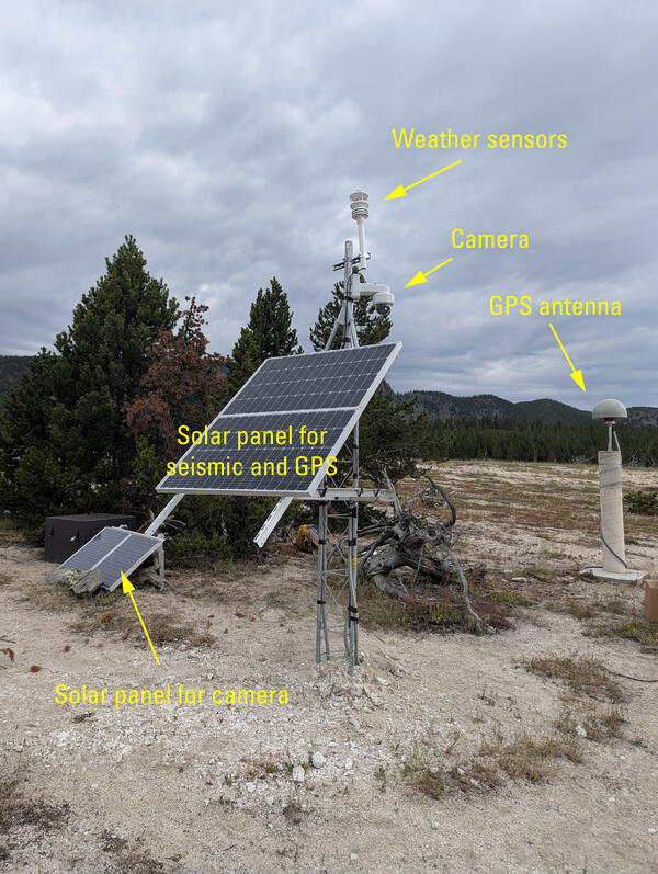 barren ground with a few small pine trees and a metal tower with a solar panel and other sensors