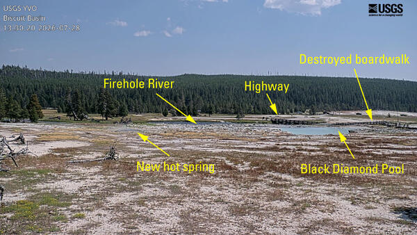 remote camera view of barren ground with a blue hot spring in the middle distance and a forested ridge in the background