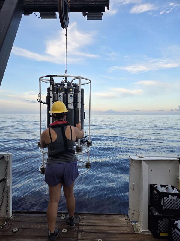 Woman in grey shirt and purple shorts with a yellow hardhat standing at the edge of the boat with the ocean in the background