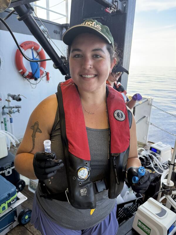 Woman in a grey shirt and red life jacket smiling at the camera while holding a few scientific collection tubes.