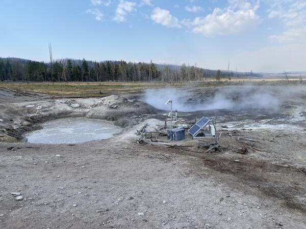 Barren ground with some equipment and solar panels in front of steaming pools.  Grassy meadow and forested area behind.