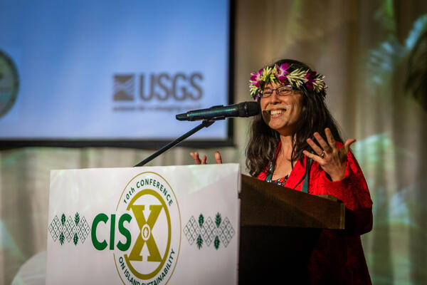 A woman with a floral crown and a red dress stands at a podium while giving a presentation at a conference.