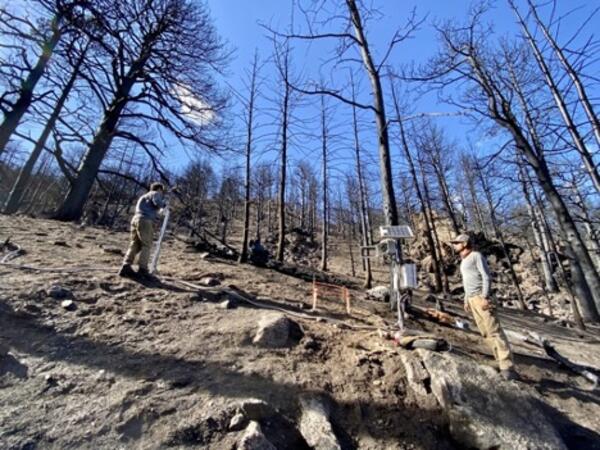 burned mountain slope with charred trees, and two people near a landslide monitoring instrument