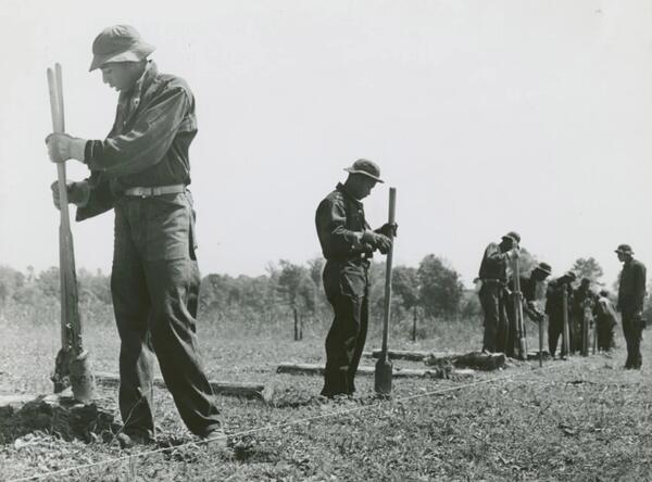 A historical image showing the Civilian Conservation Corps labor force wearing dark work overalls holding hole diggers in preparation of fence pole placement.