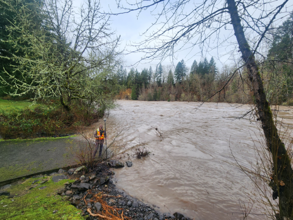 brown Clackamas River flood waters reach upper boat ramp. Drowned trees line the full river in the distance.