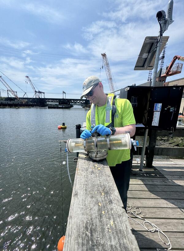 A man in a cap, neon yellow shirt, and blue gloves holds a Van Dorn sampler on a dock with a streamgage and bridge behind.