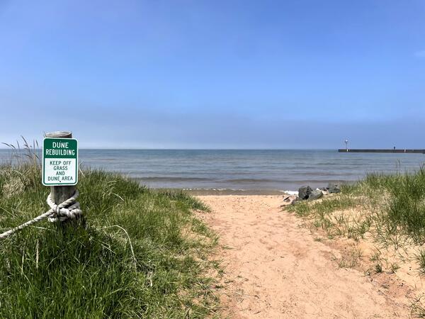 A sandy path leading to water, with a sign on the left that states "Dune Rebuilding, Keep Off Dune and Grass Area"