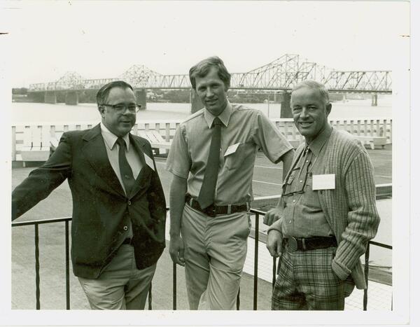 A sepia toned image shows scientists from the National Fishery Research Laboratory in 1981 standing along a metal railing with a large bridge in the background