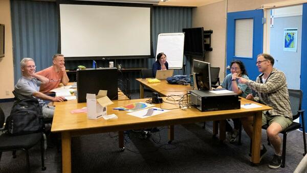 Five people sit around a large work table spread with computers and equipment