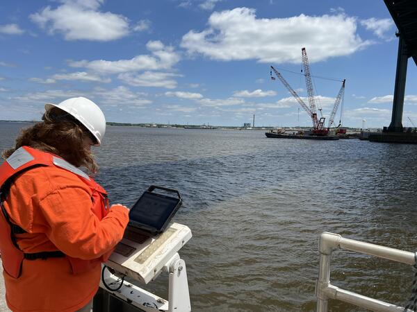 Scientist with a computer set on the bridge rail, looking out over the Delaware River at a crane barge in the distance 