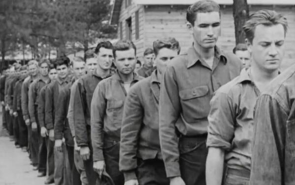 a historical image showing a line of young men in the Civilian Conservation Corps with a large wooden cabin and woods in the background