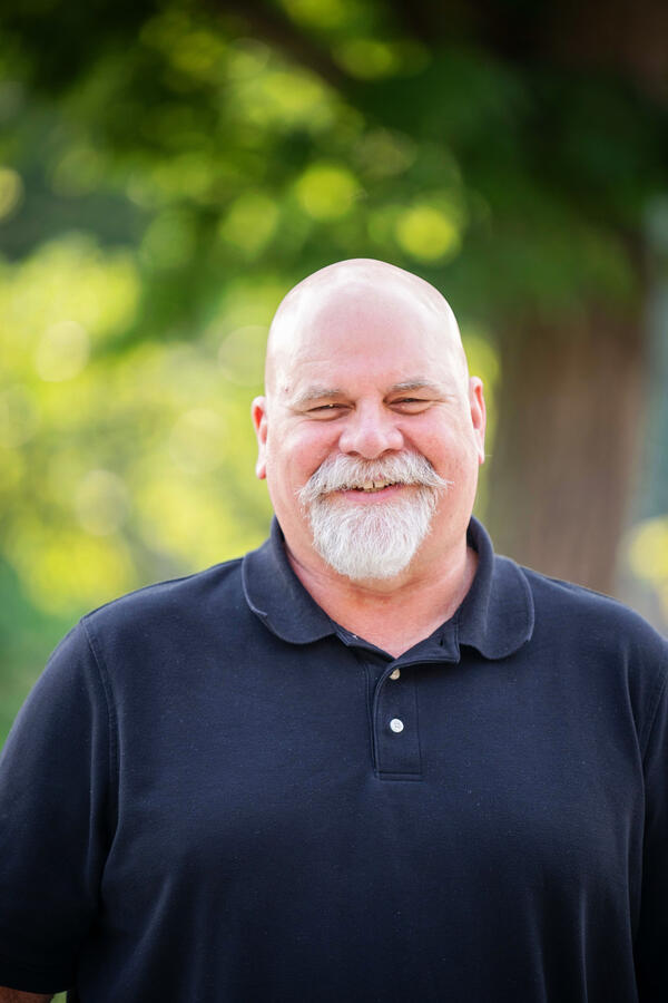 A bald, tall man with a white beard and mustache wearing a black collared shirt. 