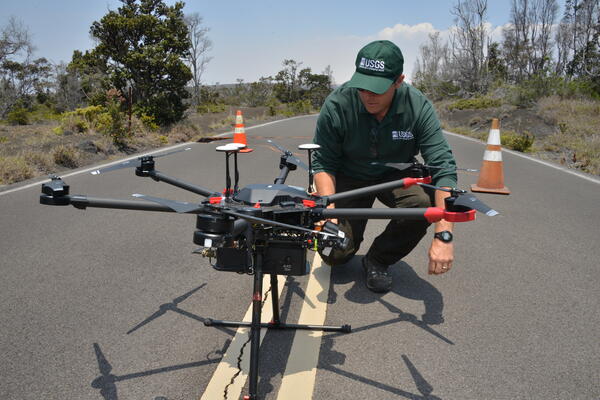 a man in dark green USGS gear kneels next to a large black drone in the middle of a road, cones and trees in the background
