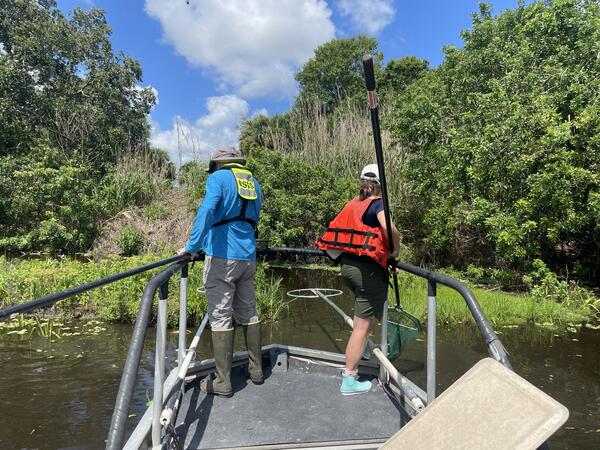 Two people in life jackets stand on a bow platform of a boat while electrofishing.
