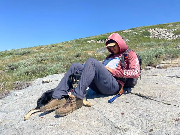 A person sitting on a large flat expanse of rock in a field. Person has a dog under their propped up knees while reading papers. 