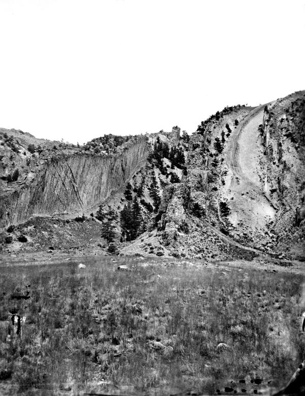 Black and white photo of a mountainside with vertical rock layers. Foreground is flat with scrubby vegetation